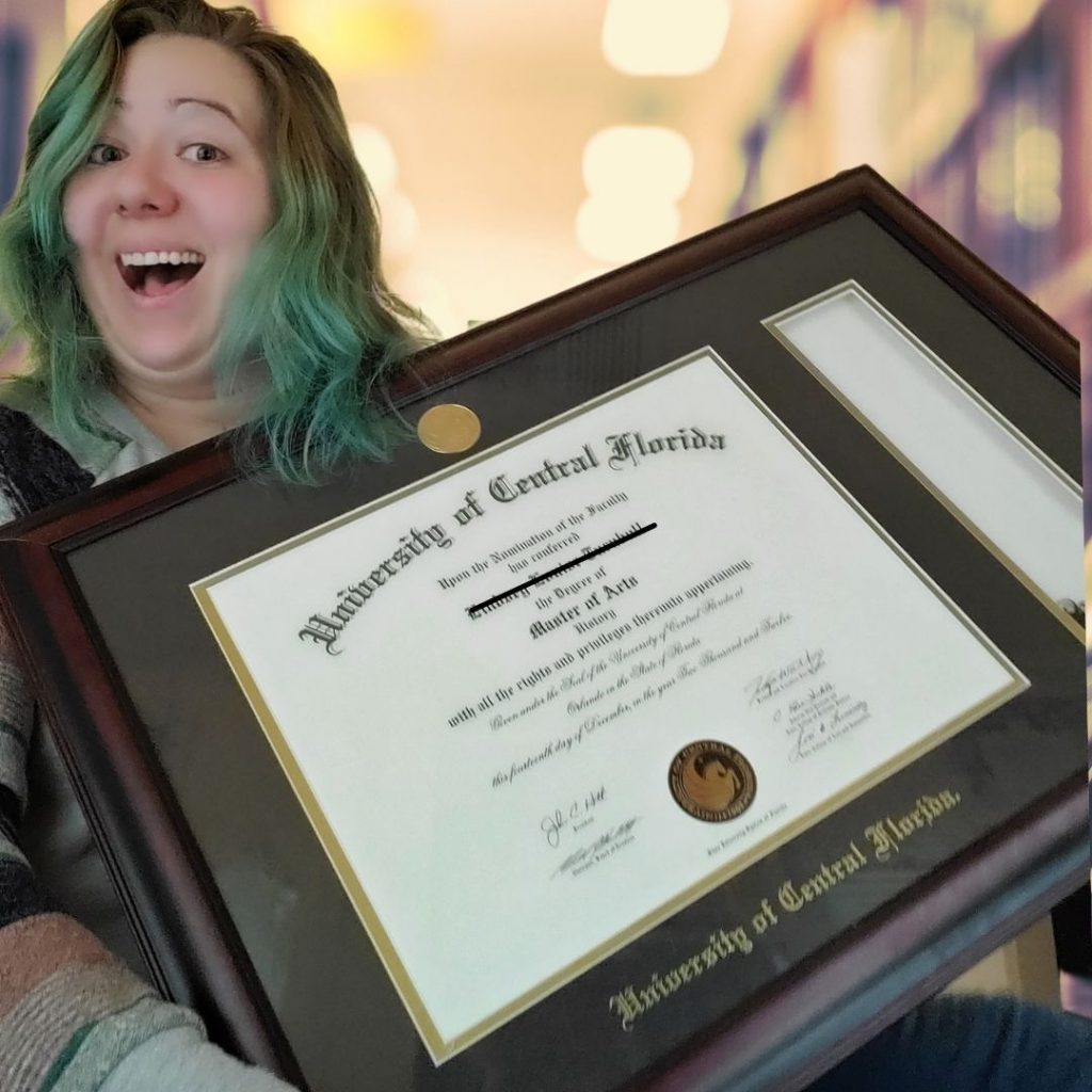 Lindsey is holding up her Master's of Arts degree in history. It covers most of her body. She is smiling and has green hair and a grey sweater.